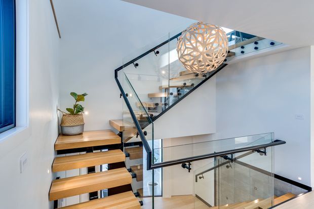 Bright contemporary stairway with floating timber treads, clear glass balustrades, slim black metal handrails, and a sculptural spherical pendant light. The stairs connect multiple levels around a light-filled void, with a small built-in timber landing shelf and potted plant.