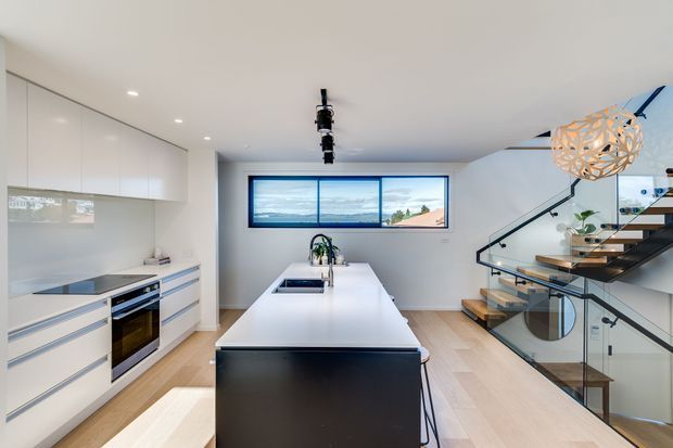 A bright contemporary kitchen with white handleless cabinetry, integrated appliances, a long white island with dark end panel, and light timber flooring. A glazed stairway with floating timber treads rises beside the kitchen, while a horizontal window provides an outlook and a sculptural pendant adds a decorative focal point.