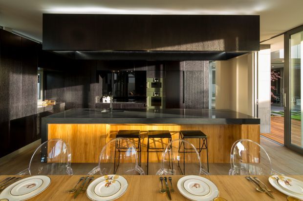 A sophisticated open-plan kitchen and dining area with a large central island, dark timber overhead cabinetry, a warm timber island face with integrated amber lighting, and a dark stone benchtop. Transparent bar stools line the island, while a timber dining table with formal place settings occupies the foreground. Full-height glazing on the right opens the room to a timber deck and brings in strong natural light.