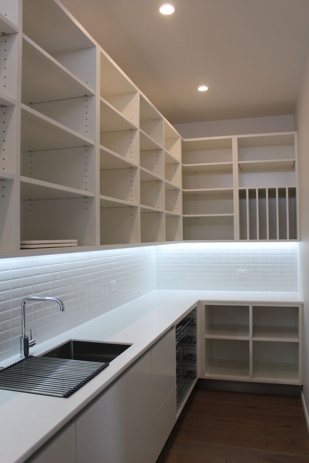 A bright, compact contemporary kitchen with extensive open white shelving, white cabinetry, pale benchtops, and a white small-format tiled splashback. The L-shaped work area includes a stainless-steel sink with drainer, integrated cooktop area, under-bench storage, and recessed ceiling lights. Dark timber flooring provides contrast.