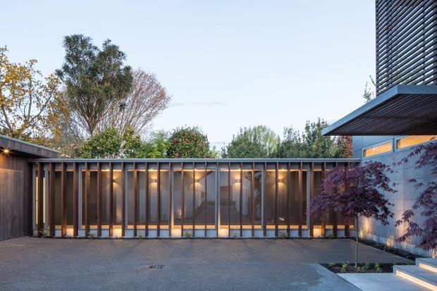 Exterior architectural view of a contemporary residential courtyard or entry area at dusk. A long glazed and timber-framed pavilion forms the main facade, with warm interior lighting visible behind vertical timber screens. Dark horizontal slatted cladding projects from the upper right building volume, while paving, integrated lighting, and a small ornamental tree define the foreground courtyard.