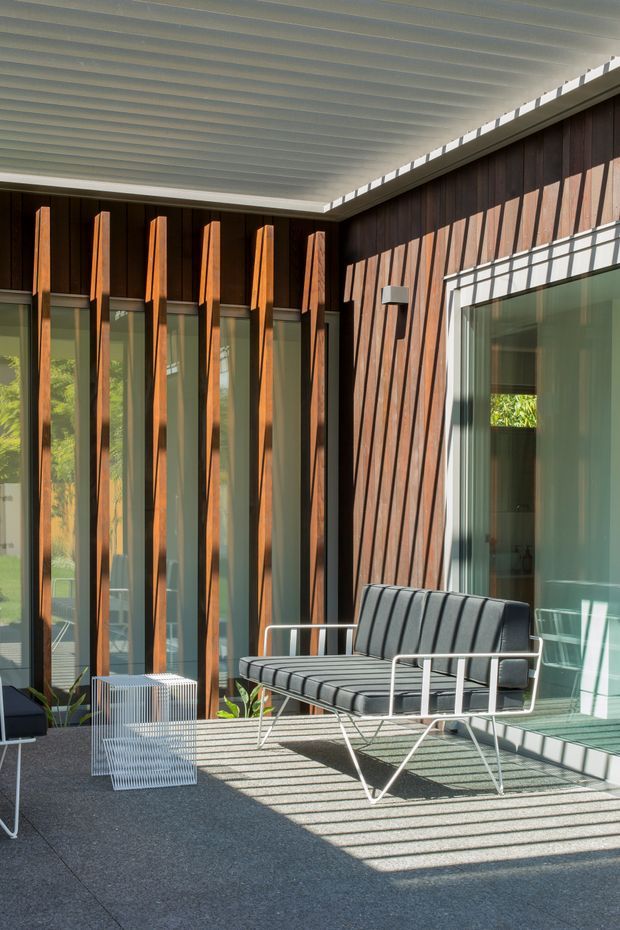 Sunlit modern outdoor terrace or covered patio featuring dark timber-clad walls, tall glazed openings with projecting vertical timber fins, a ribbed light-colored soffit, and mid-century-inspired black outdoor seating. Strong linear shadows fall across the dark paved floor, creating a refined, architectural atmosphere.