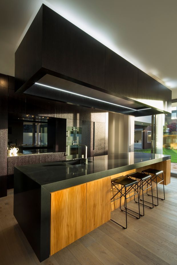 A dramatic contemporary kitchen featuring a large dark wood overhead volume with integrated linear lighting, a substantial dark stone island and benchtop, warm timber island cabinetry, black metal bar stools, and dark textured wall finishes. Full-height glazing opens the kitchen toward a bright garden or courtyard, while timber-look flooring adds warmth.