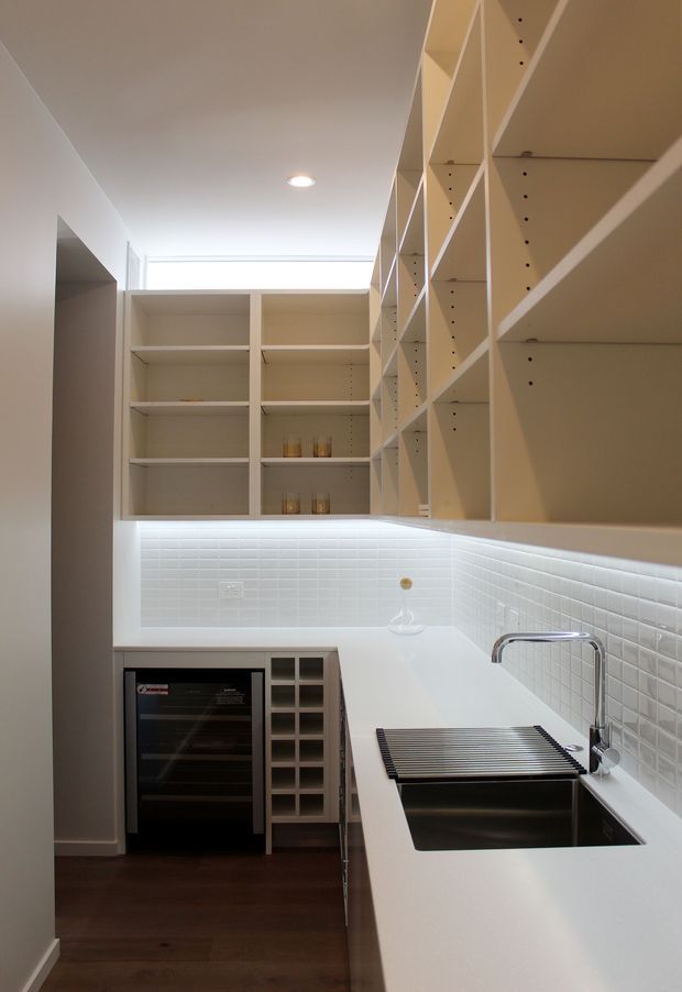 A narrow, bright contemporary galley kitchen with extensive open white shelving, white benchtops, a glossy white subway-tile splashback, stainless-steel sink and tapware, built-in oven, and dark timber flooring. A long horizontal clerestory window provides natural light above the rear cabinetry, while recessed ceiling lighting and under-shelf illumination brighten the workspace.
