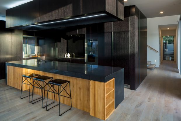 A contemporary open-plan kitchen dominated by dark vertically grained cabinetry and a large black stone island with a warm timber base. Three black metal bar stools line the island, while integrated appliances and a dark splashback create a sleek, high-contrast fit-out. A hallway and stair area are visible beyond the kitchen.