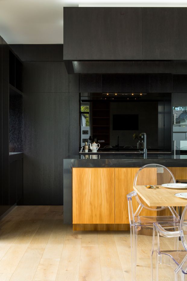 A sophisticated contemporary kitchen with dark full-height cabinetry and wall finishes, a large island featuring warm timber drawer fronts and a dark stone benchtop, and a timber dining table with transparent acrylic chairs. A mirrored or reflective backsplash area sits behind the sink, with integrated shelving and subtle ceiling lighting. Pale timber flooring contrasts with the predominantly black and charcoal fit-out.