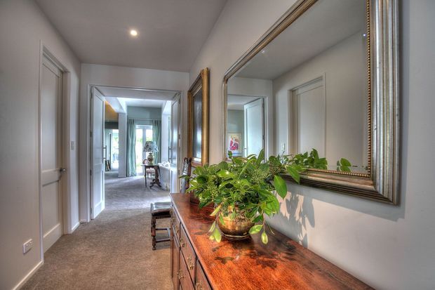 A refined residential hallway viewed from beside a long wooden console table. The corridor has light painted walls, carpeted flooring, white-trimmed doorways, recessed ceiling lighting, and large ornate framed mirrors that reflect the hallway and distant daylight. Potted greenery and decorative objects sit on the console, creating a warm, traditional interior atmosphere.
