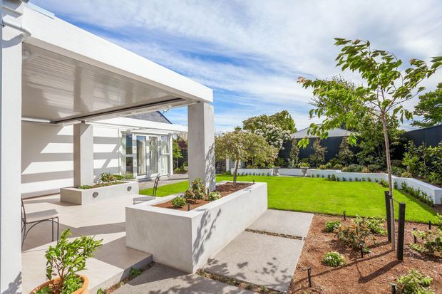 Bright contemporary residential courtyard and patio with a white flat-roofed covered outdoor area, concrete planter beds, pale paved surfaces, lawn, young trees, and dark boundary fencing. The image focuses on the designed outdoor living and garden space adjacent to the home.
