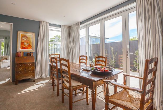 Bright traditional dining room with a solid timber dining table, matching ladder-back chairs, a wooden sideboard, framed artwork, full-height striped curtains, and extensive glazed doors and windows opening toward a fenced outdoor area.