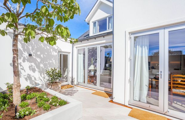 Bright exterior courtyard view of a contemporary white-painted home, featuring a raised garden bed with a mature tree, paved terrace, extensive glazed doors, and a small projecting upper-level gable window. Interior dining and living areas are visible through the glazing.