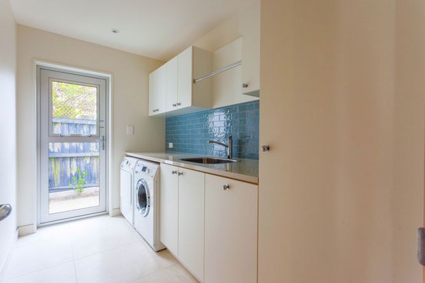 Compact, bright laundry room with white built-in cabinetry, a stainless-steel sink and benchtop, front-loading washing machine, tiled floor, and glossy blue subway-tile splashback. A glazed exterior door provides natural light and access to a small outdoor area.