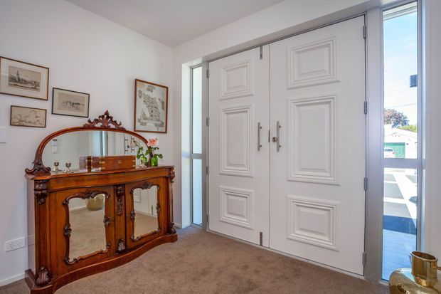 A bright, traditional entry foyer featuring a large white double front door with raised-panel detailing and glazed sidelights. An ornate dark timber console with an integrated mirror sits against the wall, accompanied by framed artwork and a small decorative plant. The floor is carpeted in a warm beige tone, and the space opens directly to an exterior entry area through the glazing.