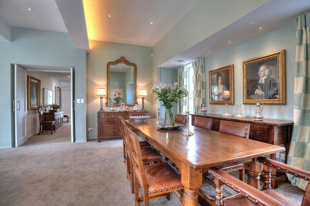 Elegant traditional dining room with a large polished timber dining table and matching leather-upholstered chairs, ornate timber sideboards, framed classical portraits, a gilded mirror, table lamps, and full-height patterned curtains. The room has soft blue-grey walls, recessed ceiling lighting, and carpeted flooring, with a hallway and adjoining rooms visible through the open doorway.