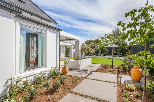 Bright exterior view of a landscaped residential courtyard beside a white rendered house. The scene includes a pitched dark shingle roof, large window, covered outdoor structure, concrete-paver walkway, raised planter walls, young trees, garden beds, and terracotta pots.
