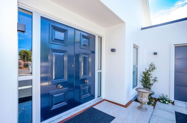 Bright contemporary residential entry courtyard featuring a prominent dark blue double front door with raised decorative panels, sidelights, white rendered walls, pale tiled paving, and a grey garage door visible to the right. A potted plant and doormat add modest entry details.