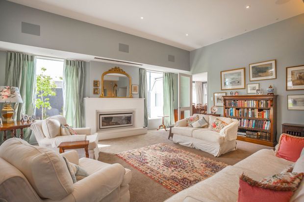 A spacious, traditionally styled living room with high ceilings, pale grey walls, large windows dressed in soft green curtains, and multiple cream upholstered sofas and armchairs. A white fireplace with a built-in firebox anchors the room, while a timber bookcase filled with books and framed artwork create a warm, refined character. A patterned Persian-style rug sits over a neutral carpet.