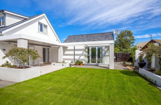 Bright exterior view of a white rendered residential home with a pitched gable roof, attached lower wing, covered patio, large glazed doors, and a neatly landscaped courtyard lawn with raised garden beds.