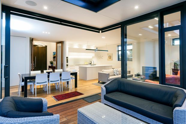 A contemporary indoor-outdoor living area viewed from a covered terrace or balcony, with dark-framed full-height glazing, a wicker outdoor seating arrangement, and a connected open-plan interior containing a dining table, compact kitchen, and living area. The interior uses light timber flooring, white cabinetry and walls, while the exterior seating is upholstered in dark blue-grey.