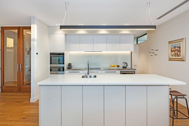 Bright contemporary kitchen with a large white island, integrated white cabinetry, pale backsplash, stainless-steel appliances, and a suspended linear pendant light. Timber parquet flooring and timber-framed glazed doors add warmth, while bar stools and artwork are visible at the right.