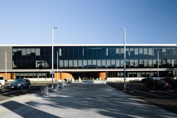 A wide exterior view of a large contemporary institutional or commercial building. The long, rectilinear facade combines extensive dark curtain-wall glazing with pale masonry or concrete panels, horizontal slab lines, and a deep covered ground-level entry. A paved forecourt with tall minimalist light poles, bollards, parking bays, and several vehicles occupies the foreground.