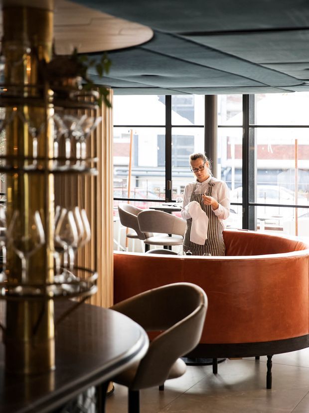 A stylish hospitality interior with a curved terracotta-red banquette, dark rounded dining chairs and tables, a large glazed window wall, and a dramatic dark segmented ceiling. A bartender stands behind the banquette, while a glassware display and vertical timber slats occupy the foreground. The space has a refined, warm, contemporary restaurant or bar atmosphere.