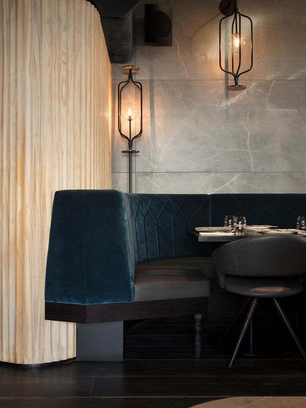 Moody, refined restaurant-style dining booth with deep blue velvet upholstery, dark timber flooring and base joinery, a softly pleated light timber wall, large grey stone-look wall panels, and sculptural black pendant lights. A small dining table and stools are visible, creating an intimate hospitality-inspired setting.