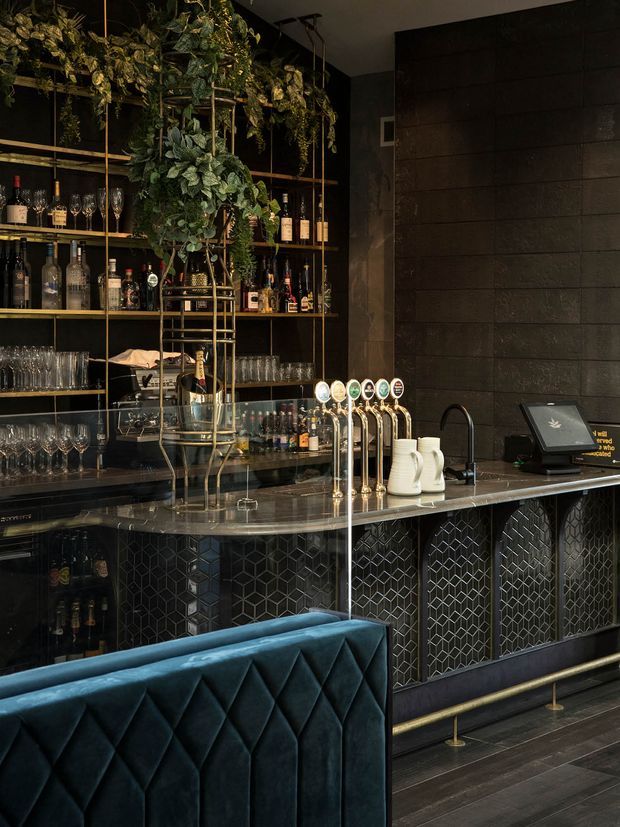 A moody, upscale bar interior with a long dark counter, multiple beer taps, a point-of-sale terminal, glassware and liquor bottles displayed on open shelving, and abundant cascading greenery. The bar front features a dark geometric patterned finish, while a deep teal quilted banquette occupies the foreground.