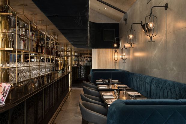 A moody, refined commercial bar and dining interior with a long brass-framed back bar and glassware display on the left, dark upholstered banquette seating and dining tables on the right, and sculptural cage-style wall sconces. The space features dark ceilings, warm metallic accents, concrete-look wall surfaces, and deep blue-green velvet upholstery.
