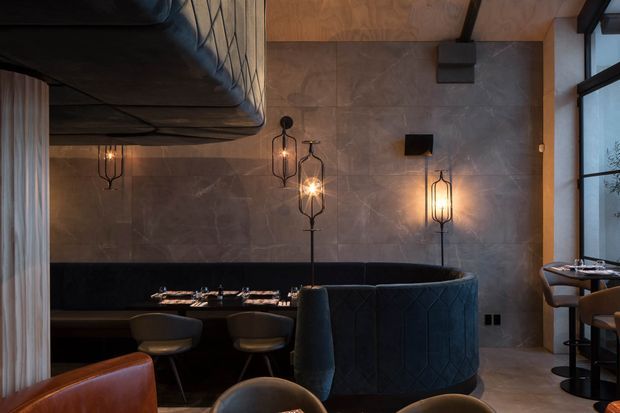 Moody upscale restaurant or hospitality interior featuring a curved upholstered booth, dark leather seating, marble-effect wall panels, warm sculptural pendant lighting, timber ceiling detailing, and large framed windows. A dining area is visible in the foreground and reflected in the booth arrangement.
