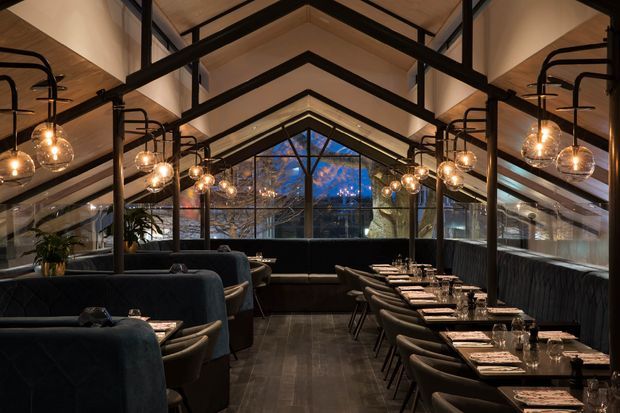 A dramatic, sophisticated restaurant interior arranged beneath a high pitched, gabled ceiling with exposed dark structural beams. Long dining tables line both sides of the central aisle, with upholstered banquette seating, dark timber-look flooring, black metal framing, and numerous warm globe pendant lights. Full-height glazing at the far end looks onto trees and evening light.