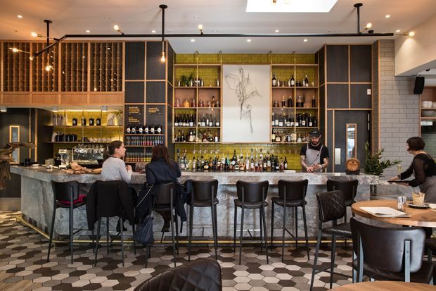 A stylish contemporary bar and restaurant interior with a long stone-topped counter, high stools, extensive built-in timber and dark-framed bottle shelving, a green tiled back bar, pendant and track lighting, and patterned geometric floor tiles. Dining tables and upholstered seating occupy the foreground, while staff and patrons use the bar.