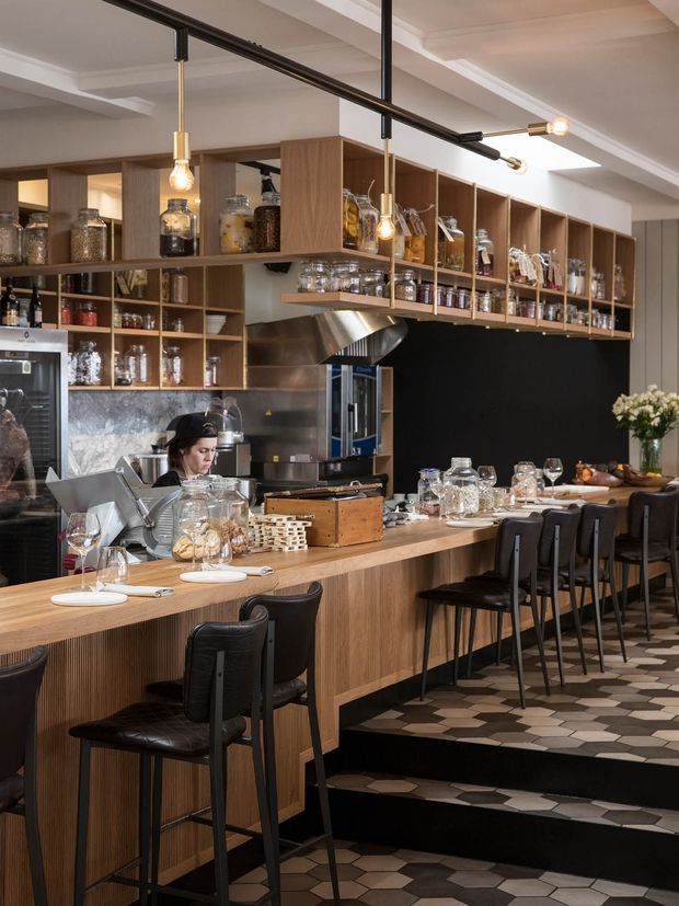 A stylish commercial hospitality interior, likely a café or restaurant, with a long timber-fronted counter and bar seating. Open timber shelving displays jars, bottles and glassware, while a commercial coffee machine, stainless-steel extraction hood and food-preparation area sit behind the counter. Black wall panels, exposed black track lighting with pendant bulbs, and a patterned hexagonal tile floor create a refined industrial-contemporary atmosphere.