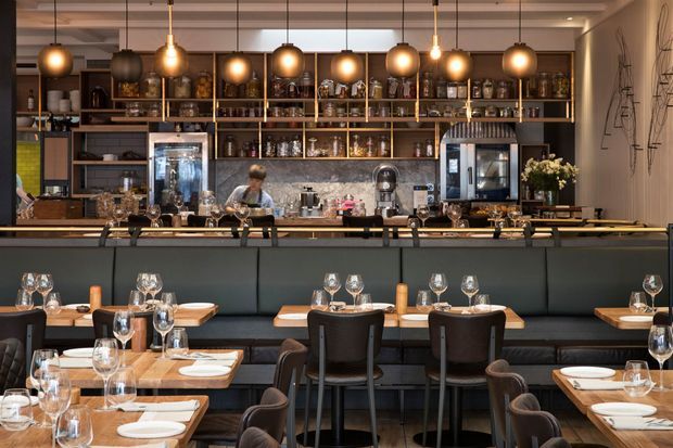 A sophisticated restaurant interior viewed across a dining area toward a long bar and open shelving. The space features dark upholstered banquette seating, timber dining tables, marble-look bar surfaces and backsplash, extensive glassware and bottle displays, and a row of warm globe pendant lights. Industrial-style artwork and dark framed glazing or partitions complete the polished hospitality setting.