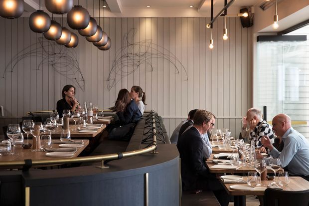 A sophisticated contemporary restaurant interior with a curved dark-toned bar or banquette in the foreground, timber dining tables, upholstered seating, and multiple diners. The space features a wall of vertical light-coloured panels with delicate linear artwork, sculptural clustered pendant lights, exposed black ceiling services, and large windows bringing in daylight.