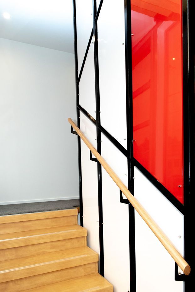 Interior view of a contemporary stairway with warm timber treads and a timber handrail. The stair balustrade features bold black metal framing with white infill panels and a striking glossy red feature panel. A carpeted landing and white walls complete the clean, graphic composition.