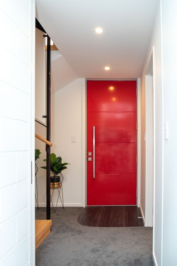 Bright residential entry hall viewed from the front entrance, featuring a bold glossy red front door with a long vertical metal handle, white walls and ceiling, recessed downlights, grey carpet, dark timber flooring at the threshold, and a partially visible stair with timber handrail and indoor plants.