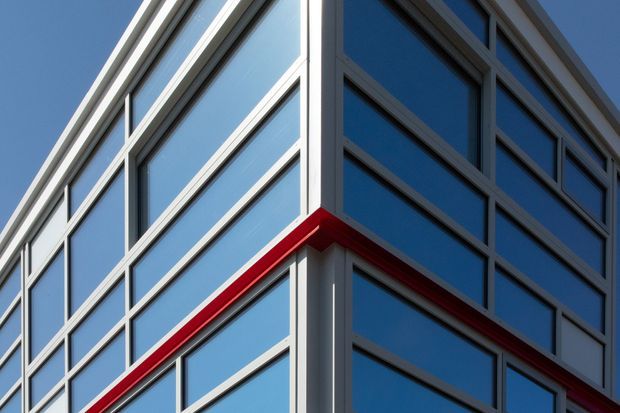 Close-up exterior view of a contemporary multi-storey building corner, featuring a gridded facade of blue reflective glazing framed by light grey metal mullions and a bold red horizontal accent band.