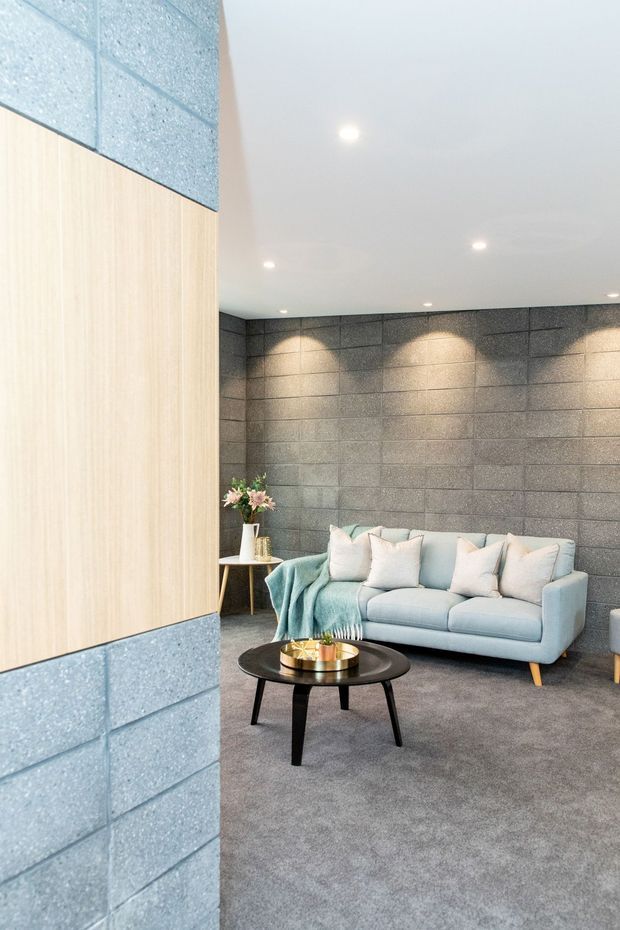 A contemporary living room with a pale blue sofa, soft cushions, a black oval coffee table, and a small side table with flowers. The room features a full-height grey concrete block feature wall with recessed spotlights, grey carpet, a white ceiling, and light timber wall panelling framed by matching blue-grey blockwork.