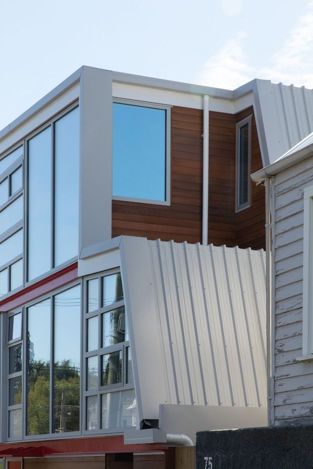 Close-up exterior view of a contemporary multi-level building addition adjoining an older weatherboard structure. The facade combines large dark-framed windows, white vertical corrugated metal cladding, warm horizontal timber wall cladding, and a small red accent band. The image focuses on the building envelope and junction between new and existing construction.