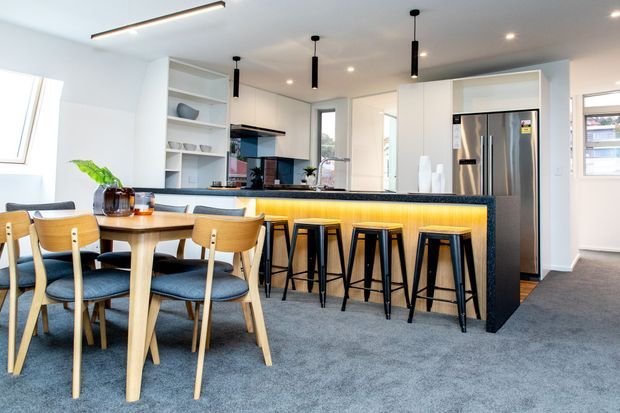 A bright open-plan kitchen and dining area with a contemporary white kitchen, dark stone-look island benchtop, warm timber breakfast bar, black pendant lights, stainless-steel refrigerator, and integrated cabinetry. A timber dining table with curved-back chairs sits in the foreground, while grey carpet and large windows create an airy, welcoming atmosphere.