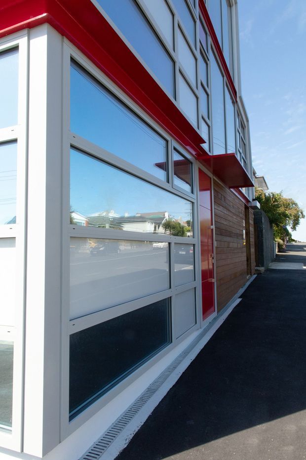Exterior close-up of a contemporary multi-storey building along a paved pedestrian edge. The facade combines extensive reflective glazing in light-coloured metal frames, bold red projecting canopy and trim elements, and horizontal timber cladding. A narrow landscaped edge and neighbouring street context are visible in the reflection and background.
