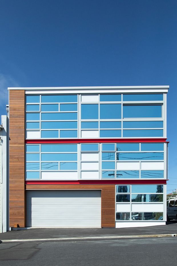 Street-facing multi-storey building facade with extensive blue-tinted grid glazing, white framing, horizontal red accent bands, and vertical timber-clad sections. The ground level includes a roller garage door and glazed entry or commercial frontage. The flat-roofed, rectilinear design has a clean contemporary appearance.