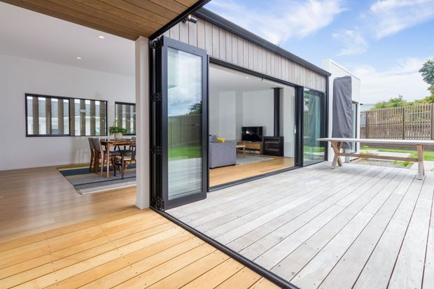 Bright contemporary indoor-outdoor living area opening onto a broad timber deck through fully folding glazed doors. The image shows an open-plan dining and living space with pale timber flooring, white walls, dark-framed glazing, and a covered timber-lined ceiling. A fenced outdoor deck with bench seating extends beyond the living room.