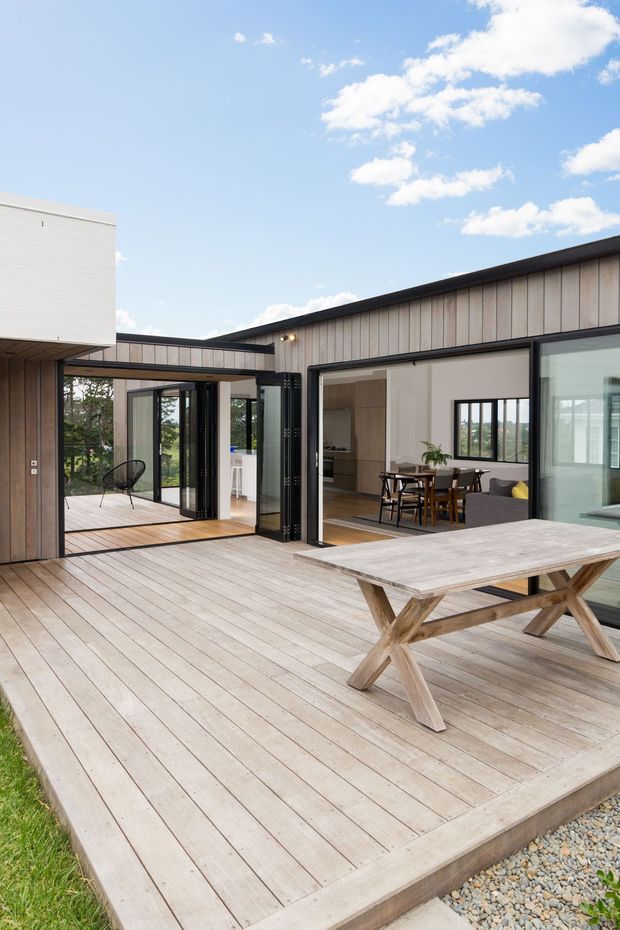 A contemporary single-storey home viewed from a large timber-clad deck. The open-plan outdoor entertaining area includes a timber picnic table, while expansive black-framed glazed doors open into a dining and kitchen area. The building has a low-pitched dark roof, vertical light timber cladding, and a covered link between wings. Bright sky and landscaped surroundings are visible beyond.