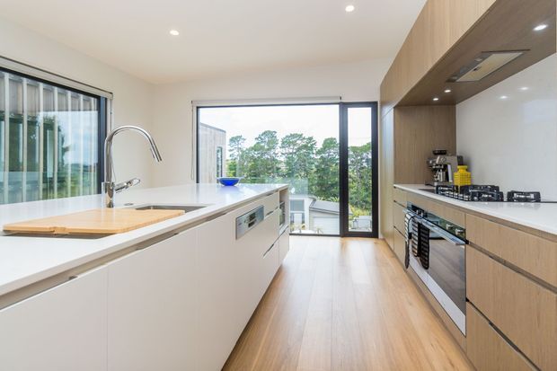 A bright contemporary kitchen with a large white island, integrated sink and curved chrome tap, timber flooring, and extensive timber cabinetry along the right wall. The kitchen includes a white benchtop, built-in oven, gas cooktop, rangehood, and full-height glazing and doors opening toward a leafy outdoor view.