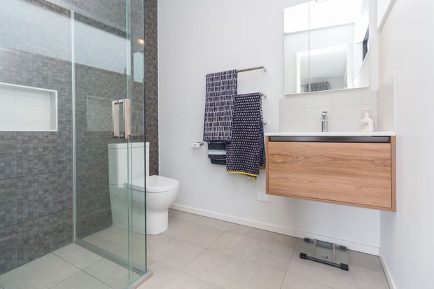 Bright contemporary bathroom featuring a frameless glass walk-in shower with grey mosaic wall tiles, a wall-hung white toilet, large-format light grey floor tiles, and a floating timber vanity with a white basin and mirrored cabinet above. Patterned towels add a dark blue accent.