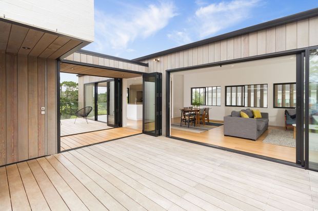 Contemporary single-level home organised around a broad timber deck and open courtyard-like outdoor living area. Large black-framed glazed openings connect the deck to a bright living and dining space, while vertical timber wall cladding and timber-lined soffits add warmth to the minimalist architectural composition.