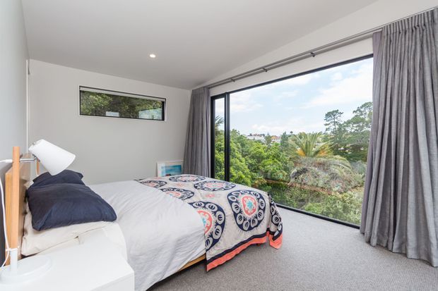 Bright contemporary bedroom with a low-profile bed, white bedding, navy pillows, and a patterned navy, white, and coral throw. Full-height sliding glass doors with grey curtains open to leafy outdoor views, while a narrow horizontal window and recessed ceiling light add to the simple, clean-lined design.