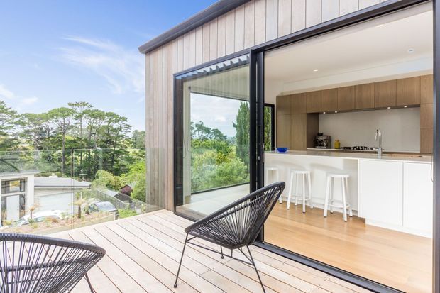 Bright contemporary kitchen opening onto a timber deck through large black-framed sliding glass doors. The kitchen features white cabinetry and island, warm timber flooring, a timber-toned splashback or rear cabinetry wall, and bar stools. A glass balustrade and leafy suburban landscape are visible beyond the deck.