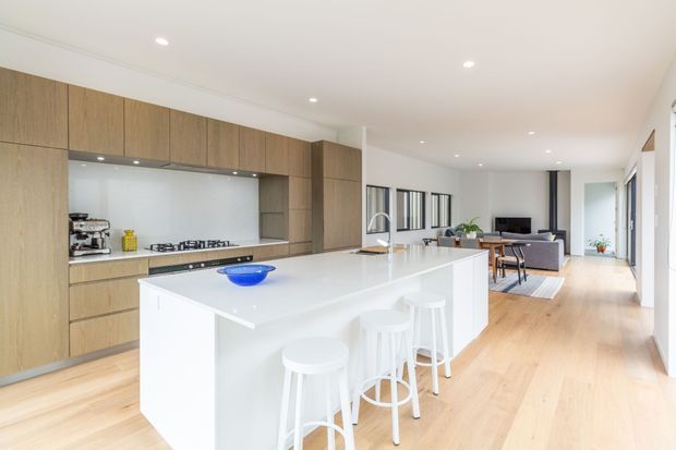 Bright open-plan contemporary kitchen with a large white island and four white stools, extensive warm timber cabinetry, a white splashback, light timber flooring, and recessed ceiling lighting. The living and dining areas are visible beyond the kitchen, with large glazed openings creating an airy connected space.